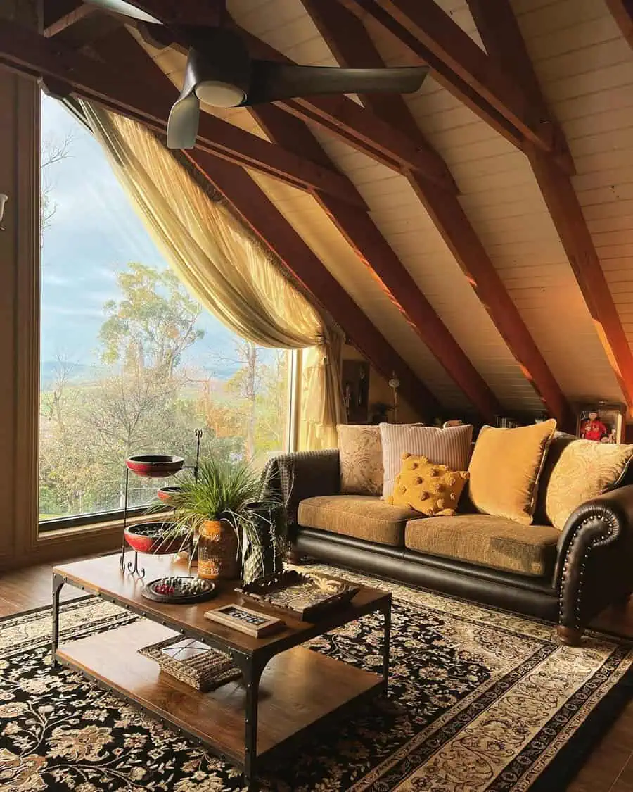 A sloped-ceiling living space with wood beams, large window, plush sofa, and a patterned rug in warm, earthy tones.