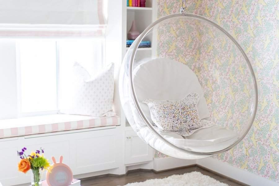 Bedroom sitting area with an acrylic swing chair, soft cushions, pastel wallpaper, and a built-in window bench for a playful and cozy feel.
