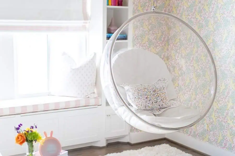 Bedroom sitting area with an acrylic swing chair, soft cushions, pastel wallpaper, and a built-in window bench for a playful and cozy feel.
