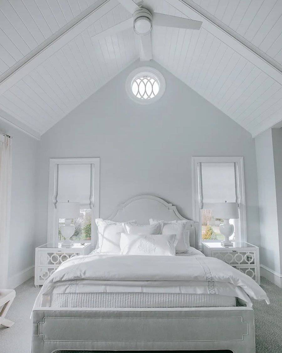 All-white bedroom with a small round window above a vaulted ceiling, featuring a white bed, ceiling fan, and matching nightstands.