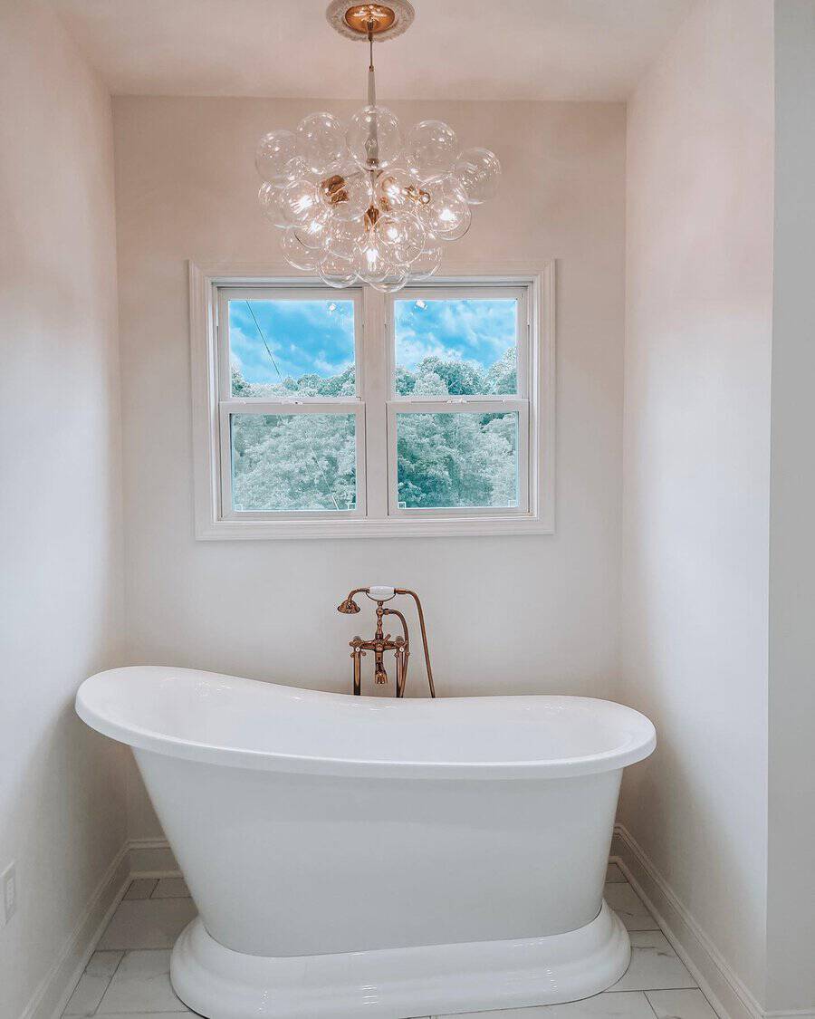 Bubble pendant above a white freestanding tub with copper faucets and a window framing outdoor greenery.