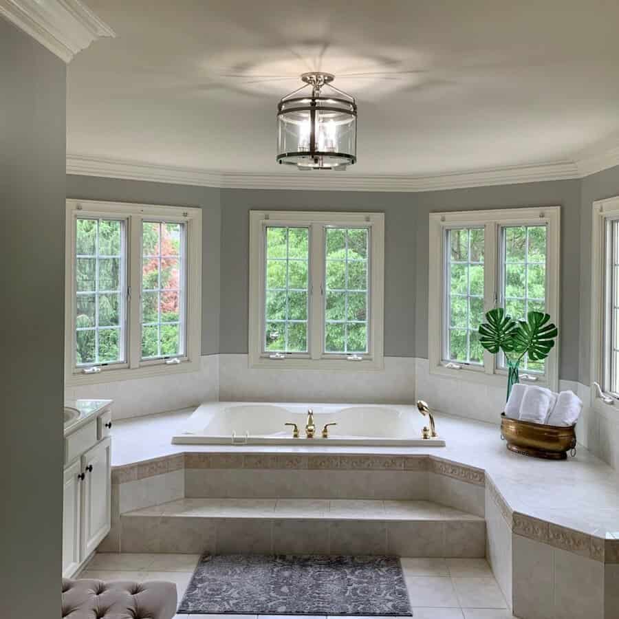 Clear drum light above an elevated corner tub with gold faucets and large windows in a tranquil gray bathroom.