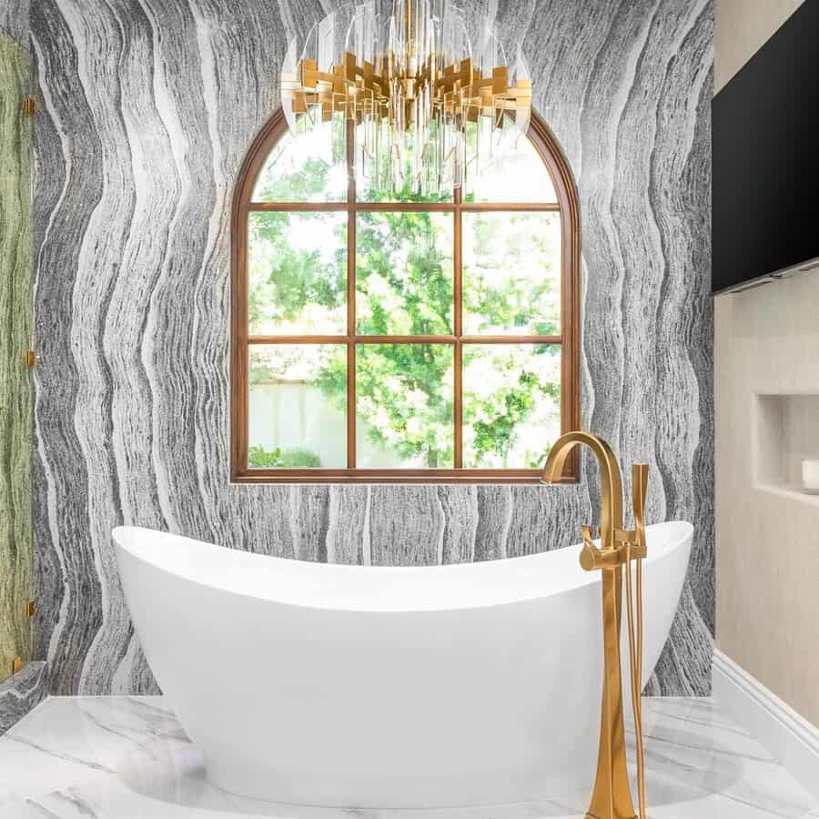 Gold-and-crystal chandelier above a sculptural white tub with swirling stone walls and an arched window.