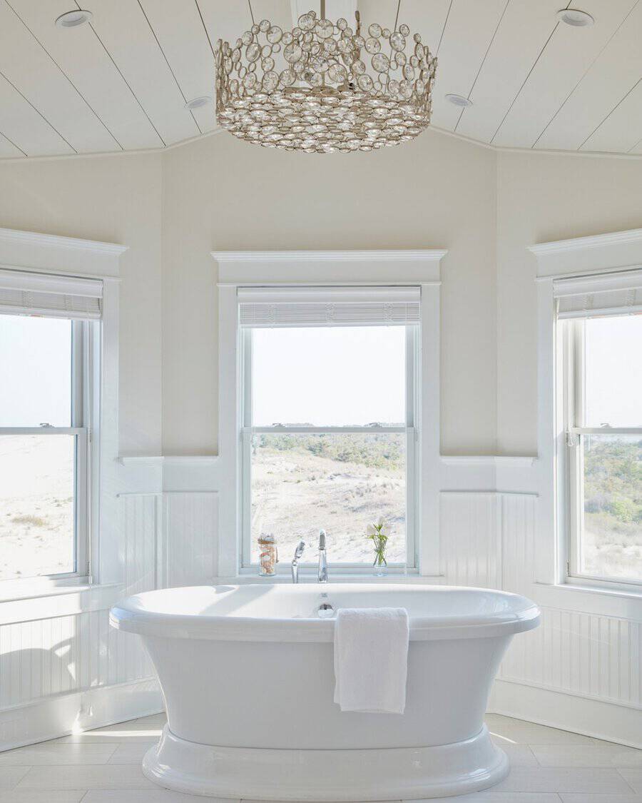 Circular disc chandelier above a freestanding tub, framed by large windows in a bright, coastal-inspired bathroom.