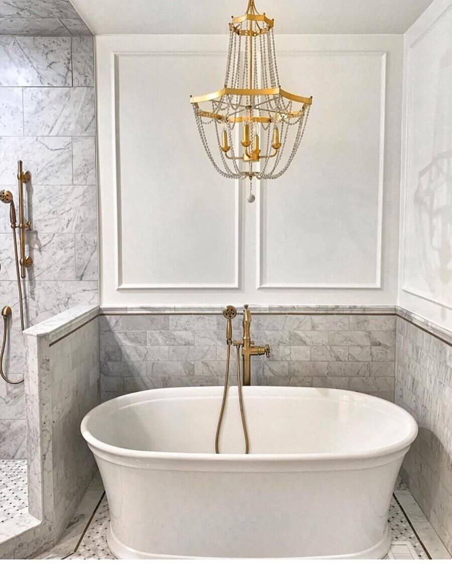 Gold tiered chandelier above a white freestanding tub in a marble-tiled bathroom with matching gold fixtures.