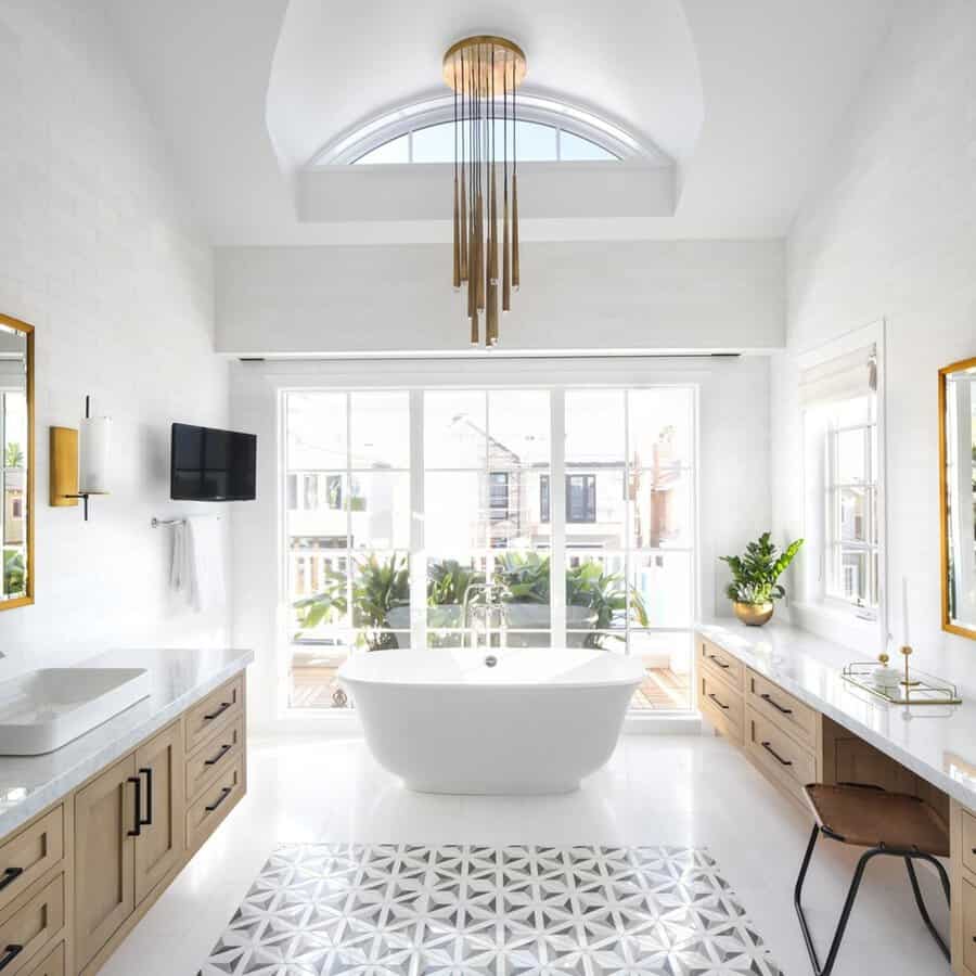 Brass multi-rod chandelier above a white tub in a bright bathroom with large windows, warm wood cabinetry, and geometric tile.