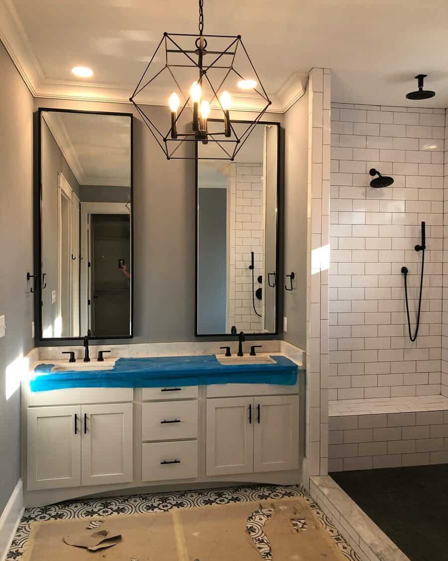 Geometric cage chandelier above a white double vanity and subway-tiled shower in a modern bathroom with black accents.