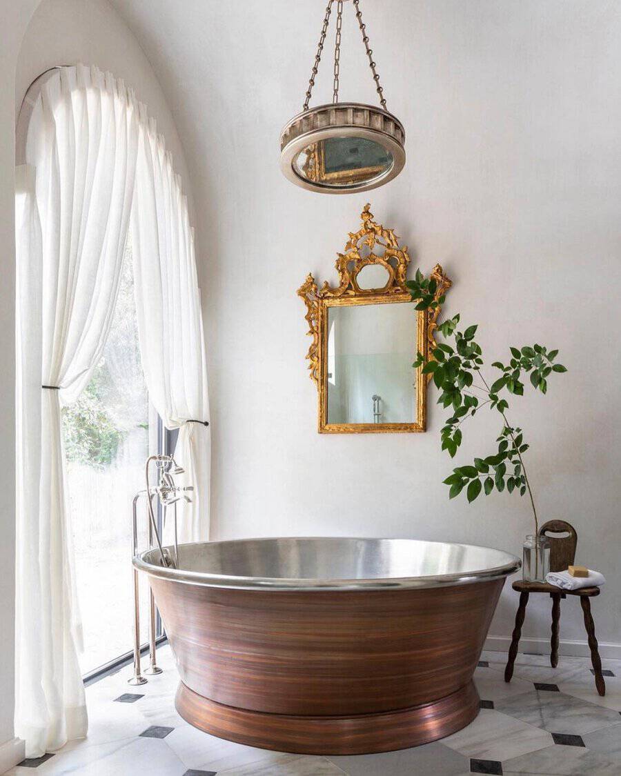 Round chandelier over a copper soaking tub with a gilded mirror, sheer drapery, and marble flooring in a bright bathroom.