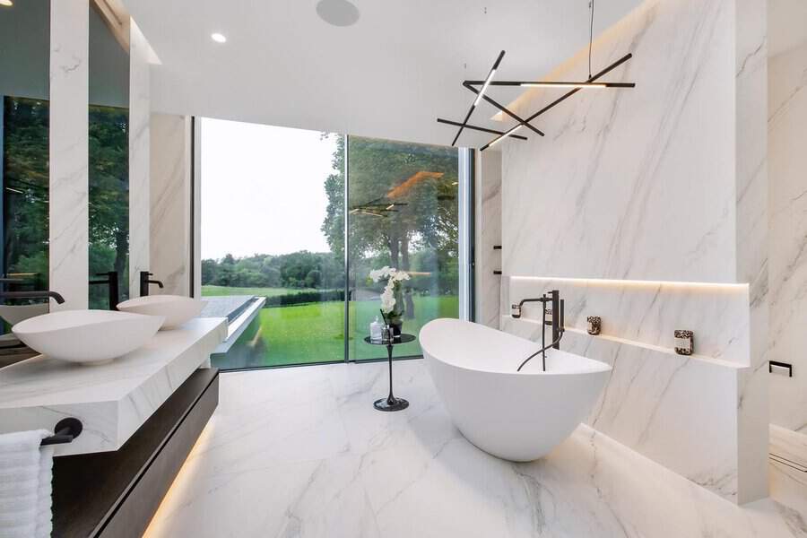 Minimal LED rod fixture above a freestanding tub, large glass wall, and marble surfaces in a modern bathroom setting.