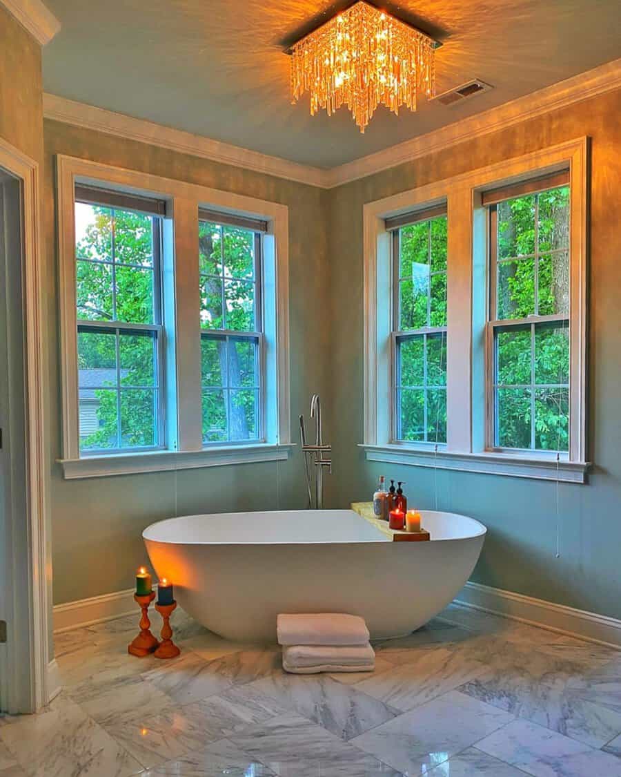 Square crystal chandelier above a freestanding tub in a soft green bathroom with large windows and marble flooring.