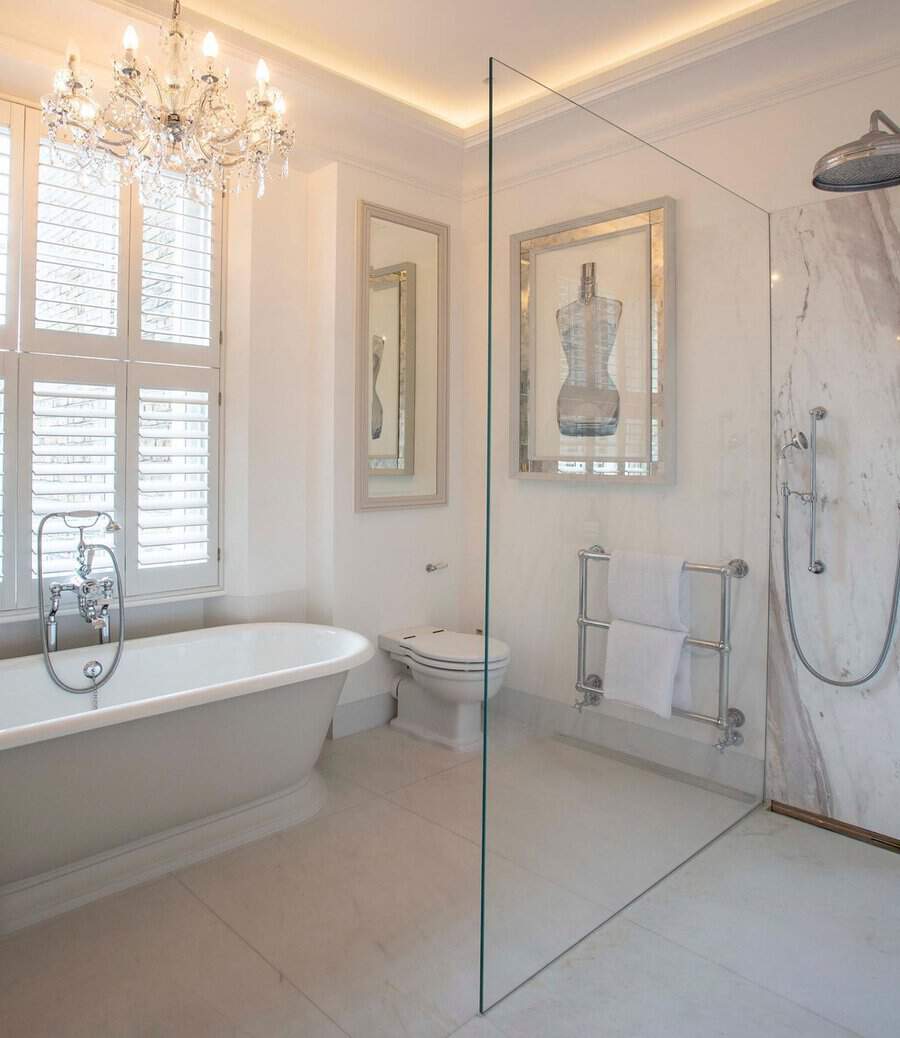 Maria Theresa chandelier with glass arms above a white tub in a bright, neutral-toned bathroom with shutters.