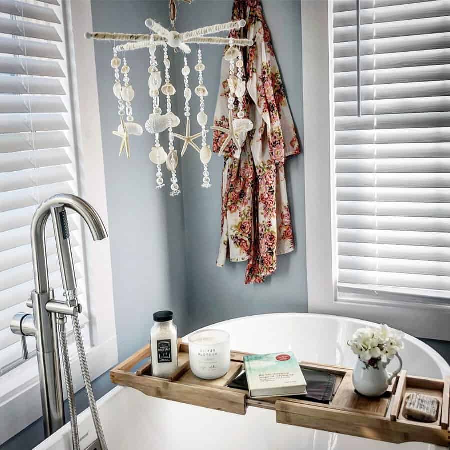 Shell mobile above a white freestanding tub with a wooden caddy, floral robe, and beachy décor accents.