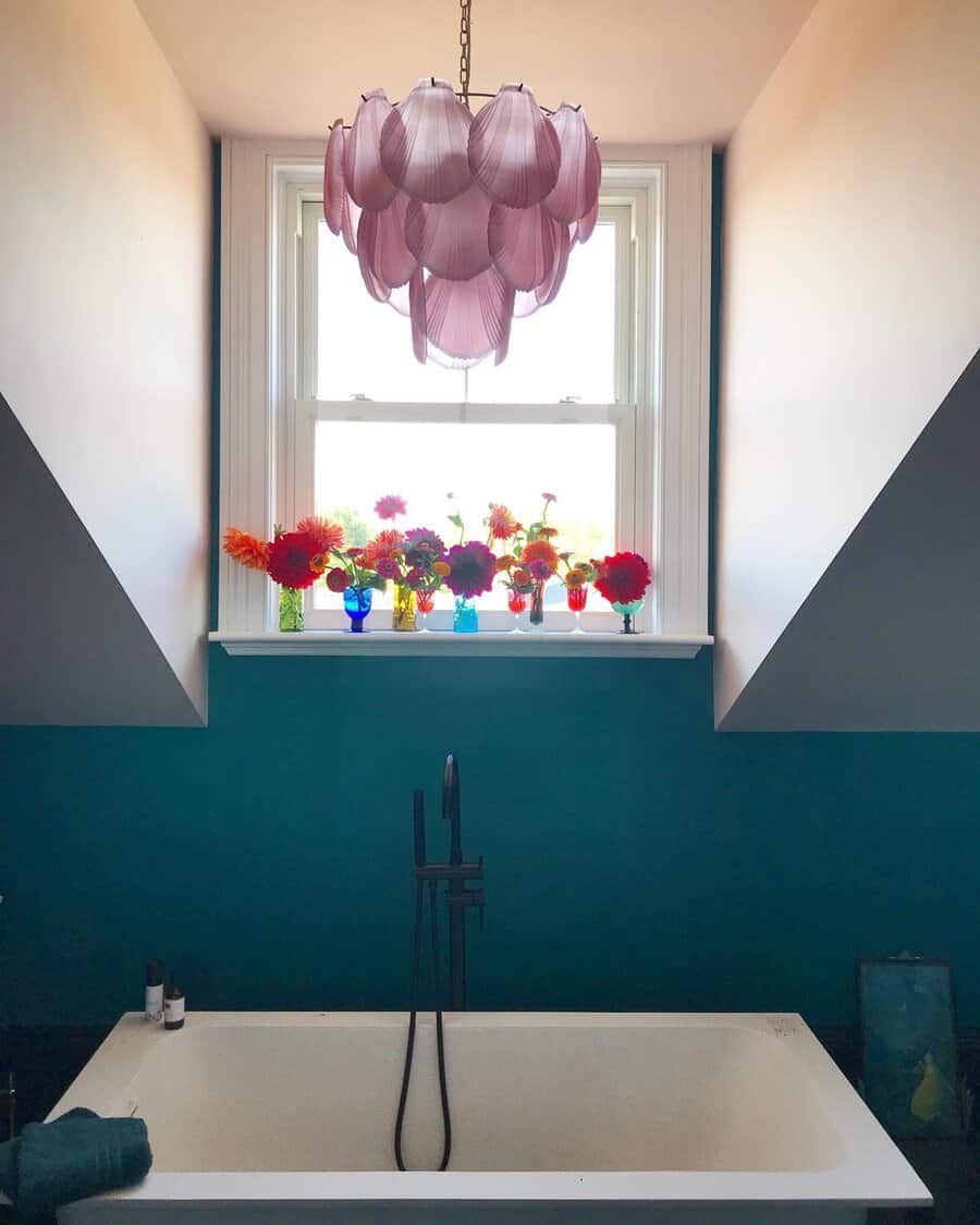 Layered pink pendant above a white tub with teal walls, framed by bright blooms on a windowsill in colorful vases.