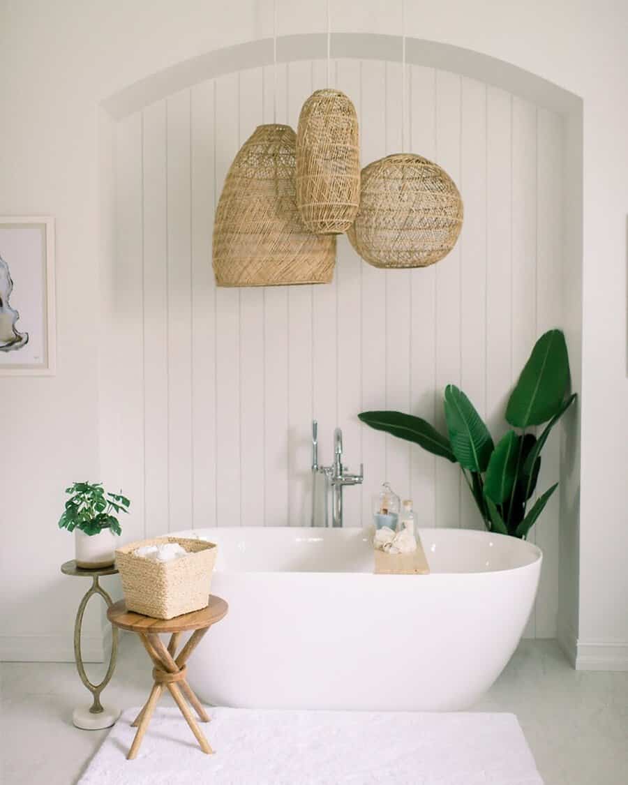 Wicker pendant lights above a white freestanding tub framed by shiplap walls, a wooden stool, and a leafy plant.