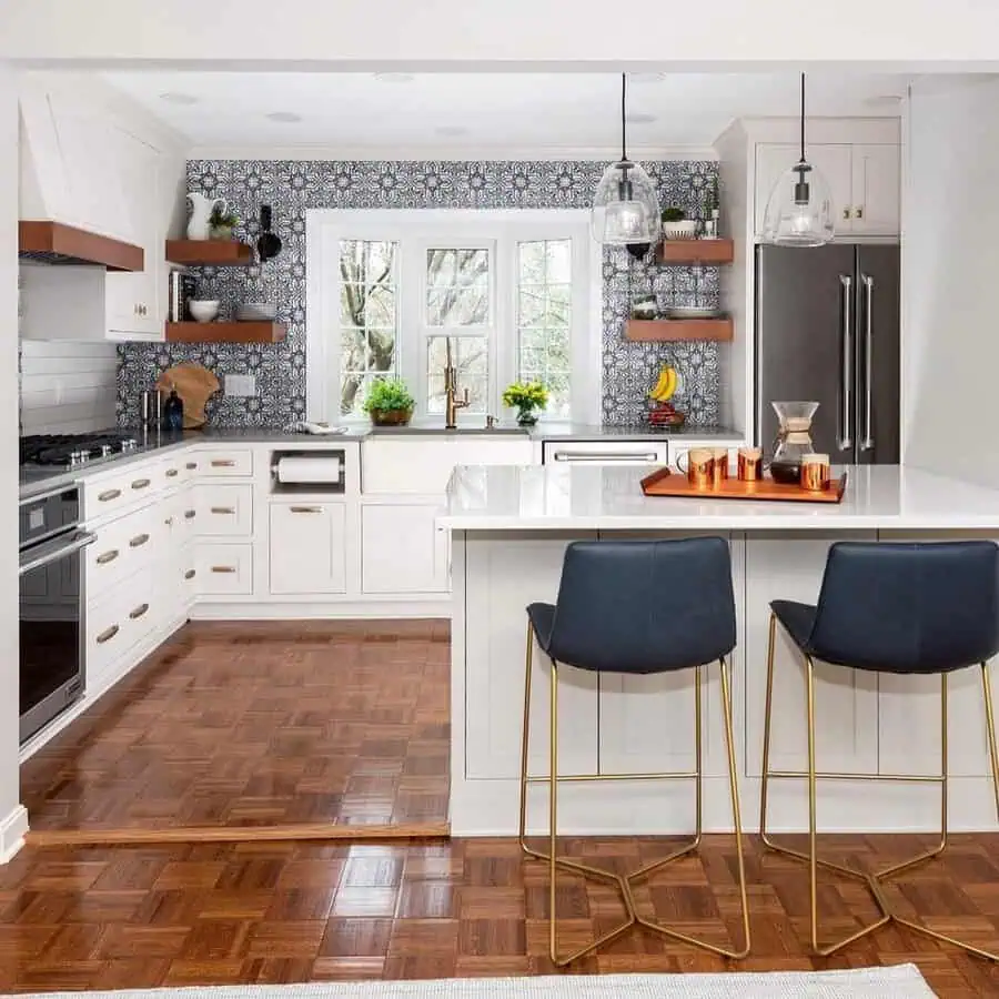 White kitchen with patterned backsplash, bay window, floating shelves, gold hardware, and navy bar stools.