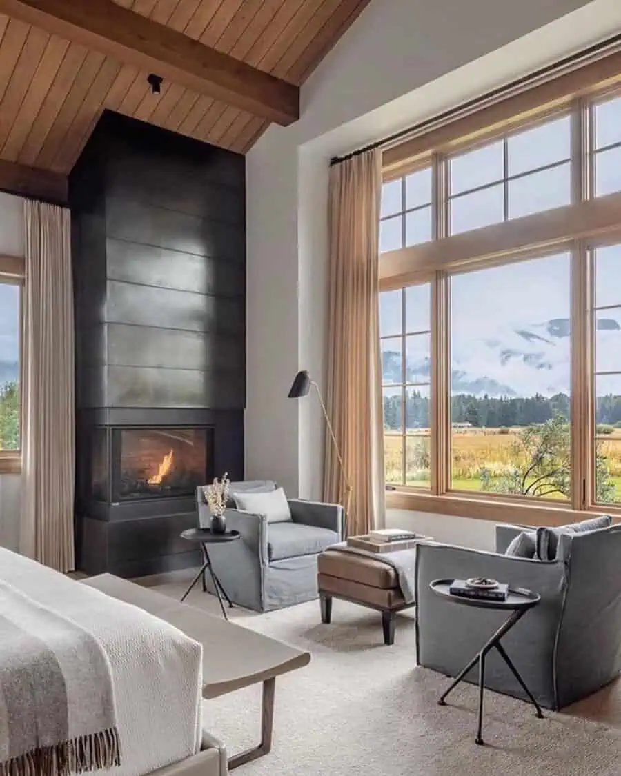 Bedroom sitting area with a fireplace, large windows, neutral armchairs, wood accents, and soft textiles for a cozy retreat.