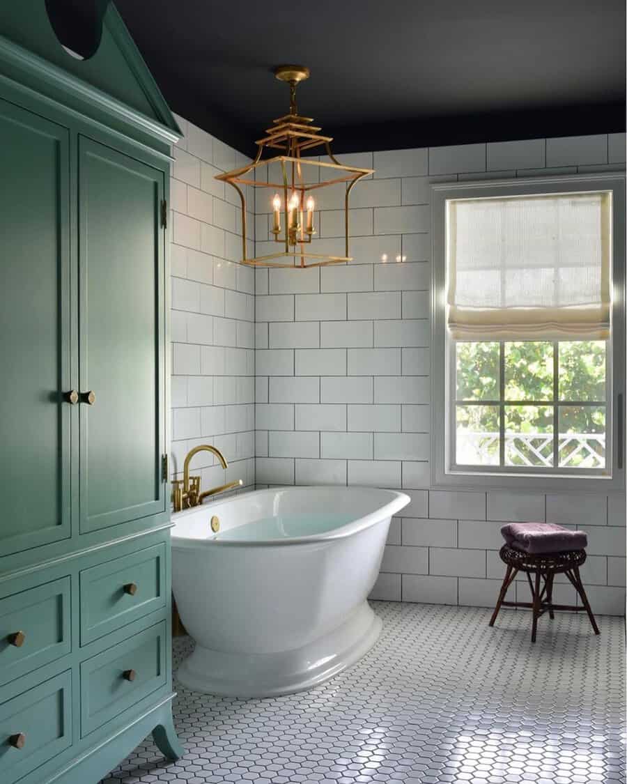 Brass lantern chandelier above a freestanding tub, white subway tile, and a green armoire in a stylish bathroom.