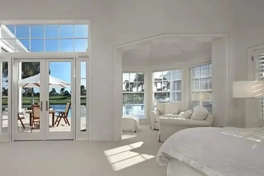 Bright bedroom sitting area with large windows, white furniture, a cozy armchair, and an outdoor view.