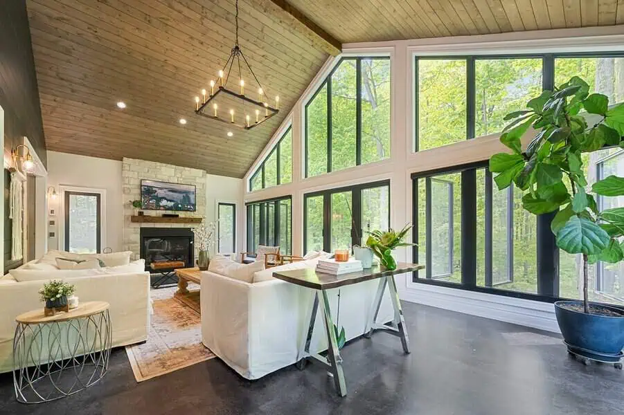 Spacious living room with a tall wood ceiling, stone fireplace, and huge windows offering forest views, furnished in soft neutrals.