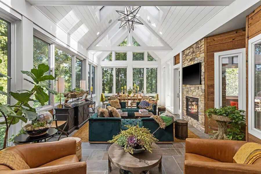 Sunlit living and dining space with a white cathedral ceiling, star chandelier, big windows, plush seating, and a stone fireplace.