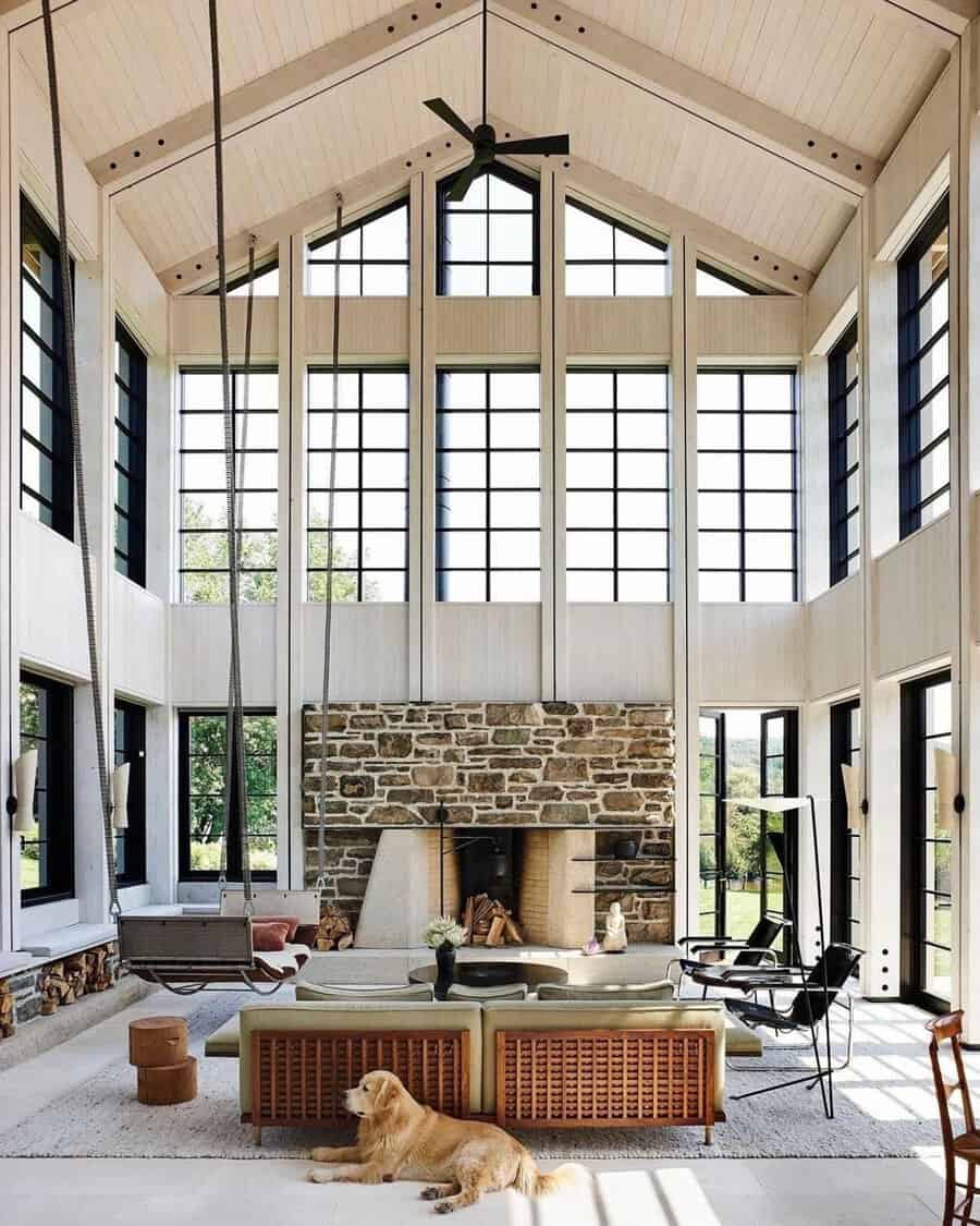 Light-filled space with soaring ceiling, stone fireplace, black-framed windows, and a dog near a suspended seat.
