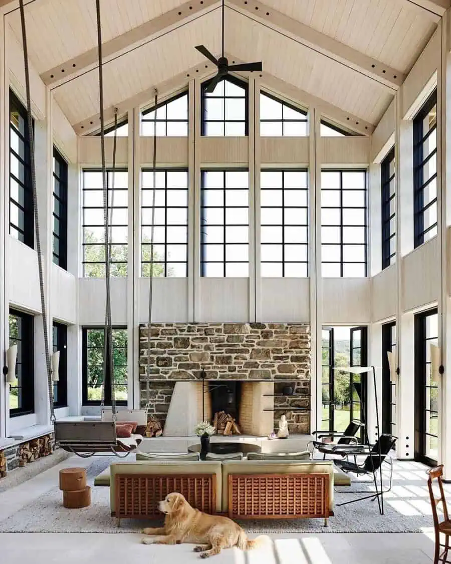 Light-filled space with soaring ceiling, stone fireplace, black-framed windows, and a dog near a suspended seat.