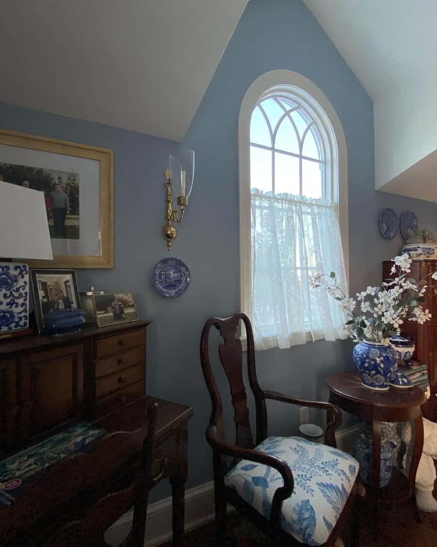 Classic bedroom sitting area with a vintage chair, blue walls, lace curtains, antique furniture, and decorative plates.