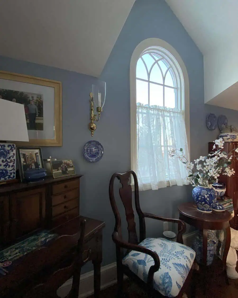 Classic bedroom sitting area with a vintage chair, blue walls, lace curtains, antique furniture, and decorative plates.