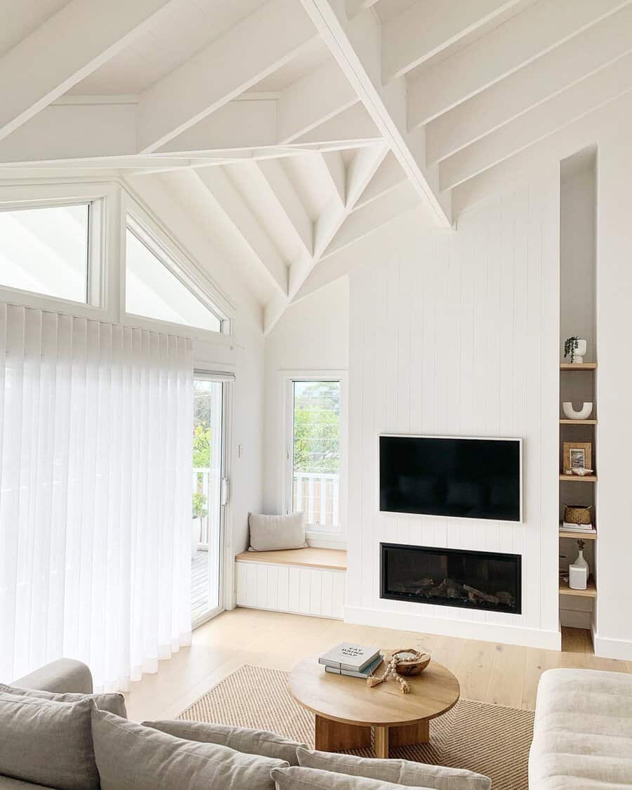 Bright living room with white cathedral ceiling, exposed beams, a built-in window seat, and a modern fireplace below a mounted TV.