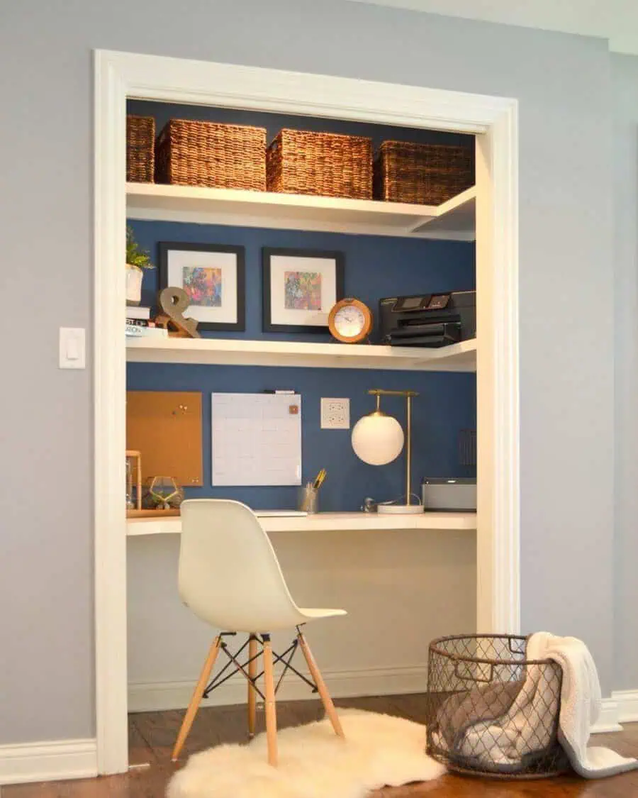 Closet office with a blue accent wall, floating shelves, woven storage baskets, a built-in desk, modern lighting, and a white mid-century chair.
