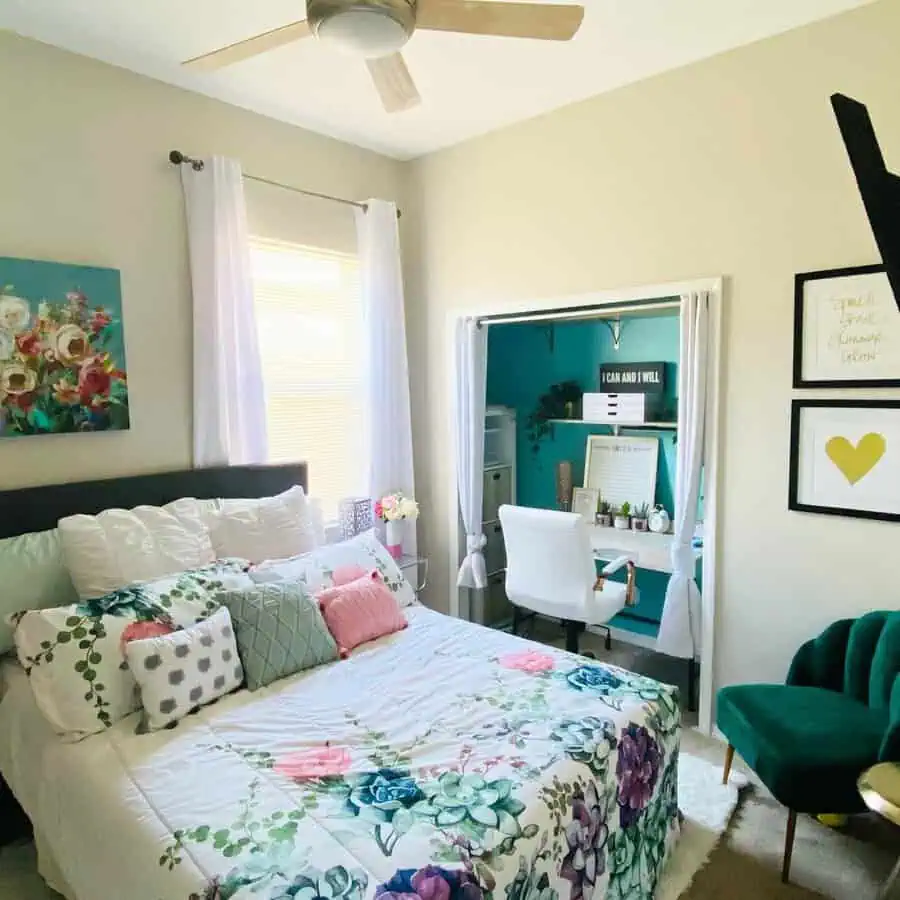 Closet office in a bedroom with a teal accent wall, white desk, floral bedding, cozy seating, and sheer curtains for a flexible setup.
