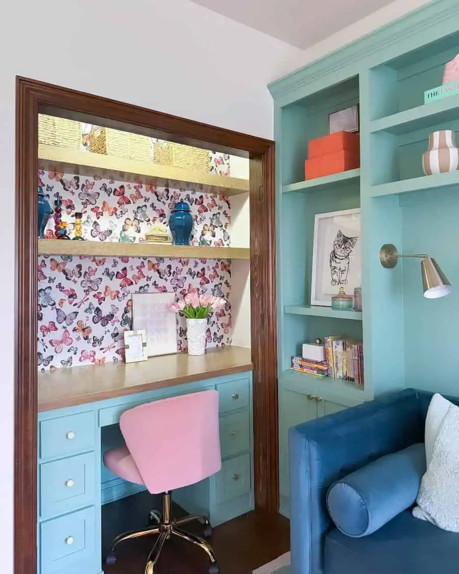 Closet office with butterfly wallpaper, turquoise built-in desk, pink chair, wood shelving, and colorful decorative accents.