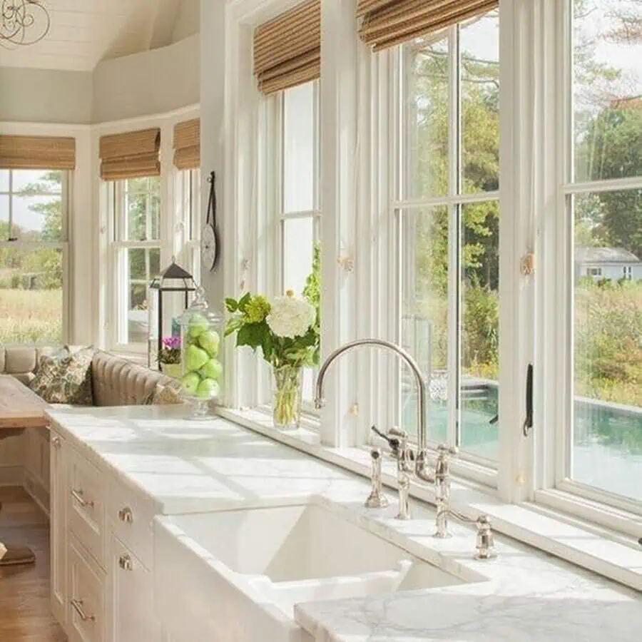 White kitchen with tall windows, marble countertops, and a comfy banquette looking out onto greenery.