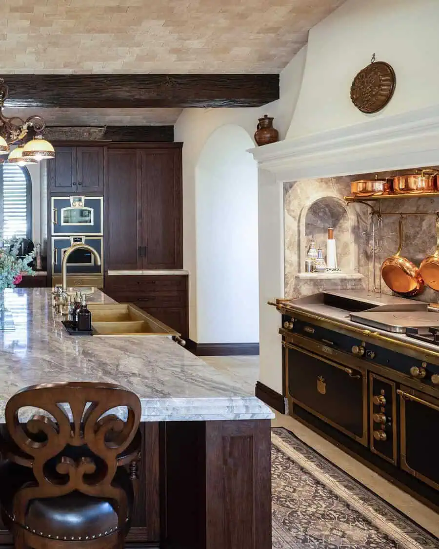 Spanish-style kitchen with veined stone countertops, dark wood cabinetry, a black and brass range, and an arched cooking alcove with copper pots.