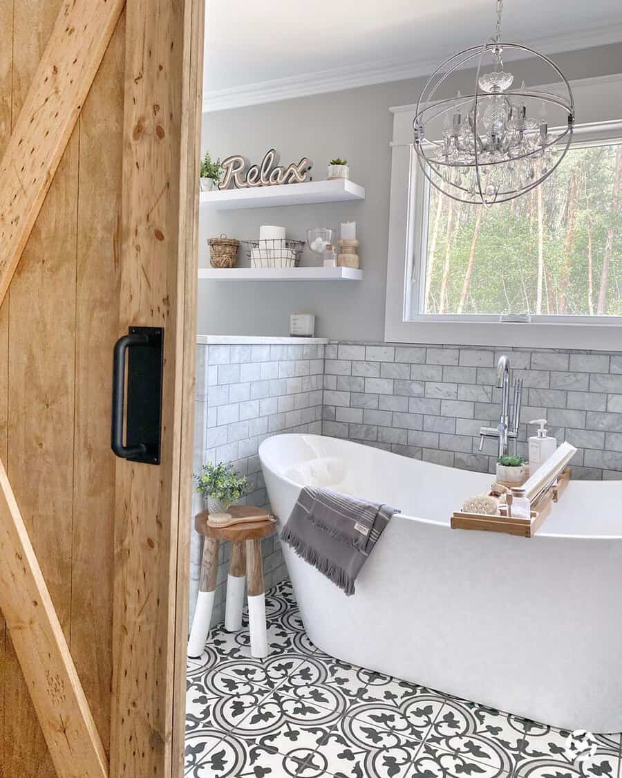 Orb chandelier above a freestanding tub, barn door, and patterned tiles in a farmhouse-inspired bathroom.