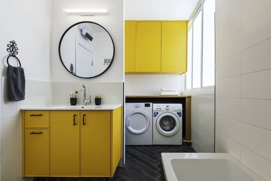 Bathroom-laundry area with bright yellow cabinets, a round black mirror, white appliances, and dark herringbone flooring.