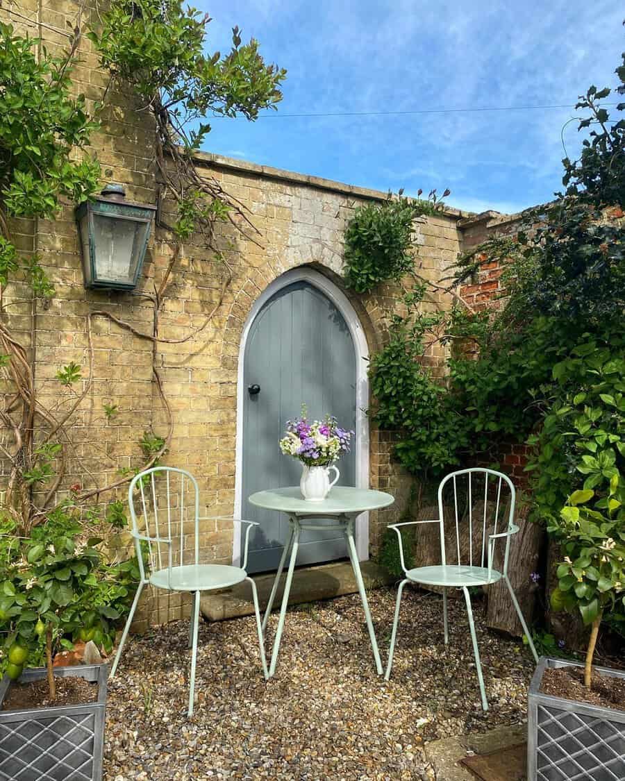 Pastel bistro set before a grey arched door in a brick courtyard, surrounded by greenery under a bright blue sky.