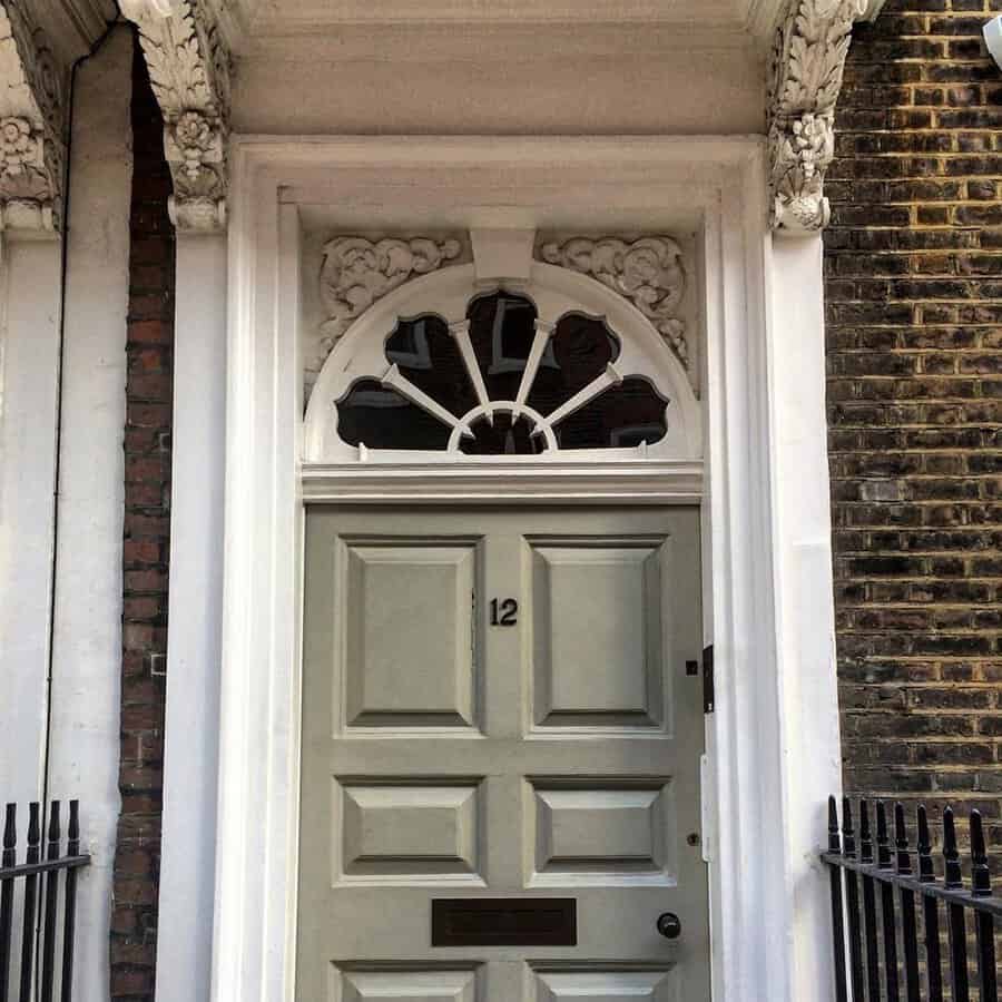 Georgian front door in a neutral shade, framed by an ornate fanlight and carved detailing against a brick facade.