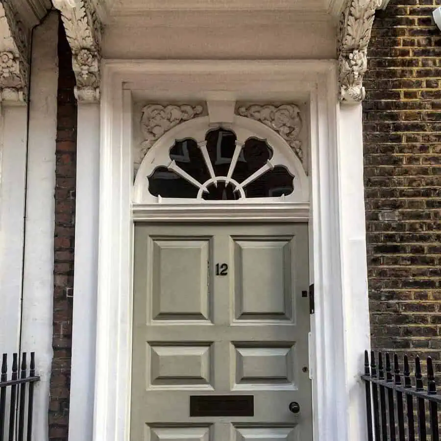 Georgian front door in a neutral shade, framed by an ornate fanlight and carved detailing against a brick facade.