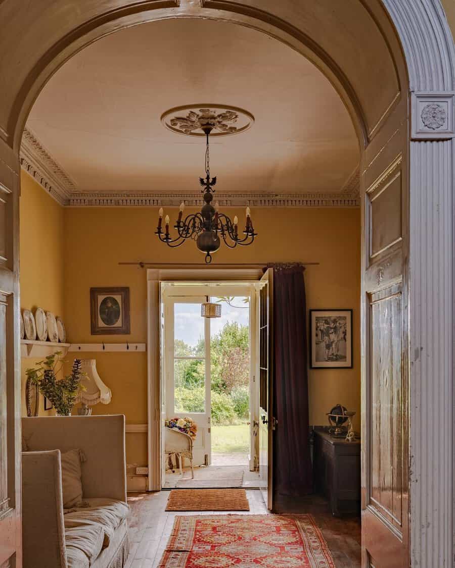 Georgian hallway with arched doorway, yellow walls, ornate chandelier, and an open door revealing a garden view.