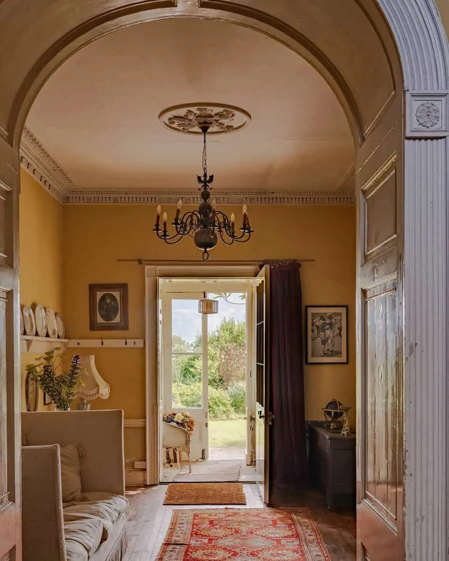 Georgian hallway with arched doorway, yellow walls, ornate chandelier, and an open door revealing a garden view.