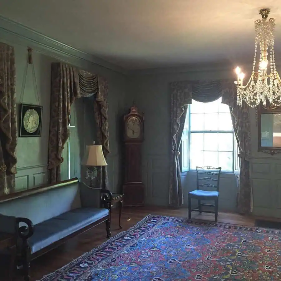 Georgian drawing room with green paneled walls, a grandfather clock, ornate chandelier, and plush drapery.