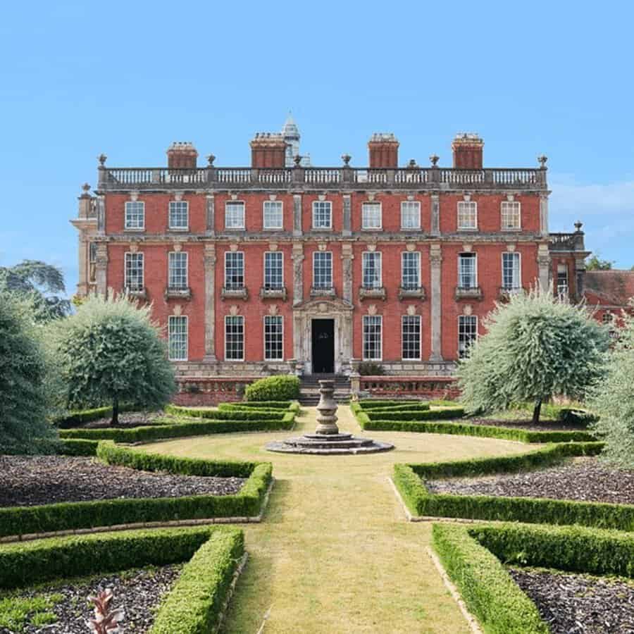 Formal garden with hedges and a central fountain leading to a large red-brick Georgian house under a bright blue sky.