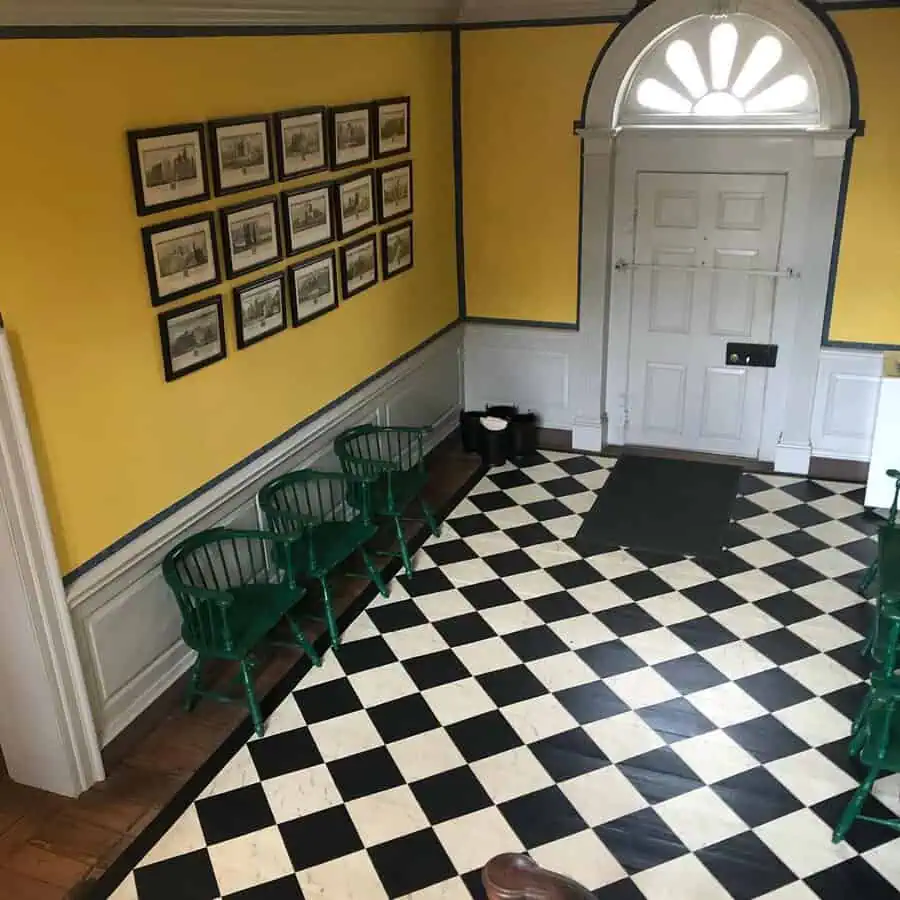 Yellow Georgian foyer with fanlight door, checkerboard floor, and green Windsor chairs against paneled wainscoting.