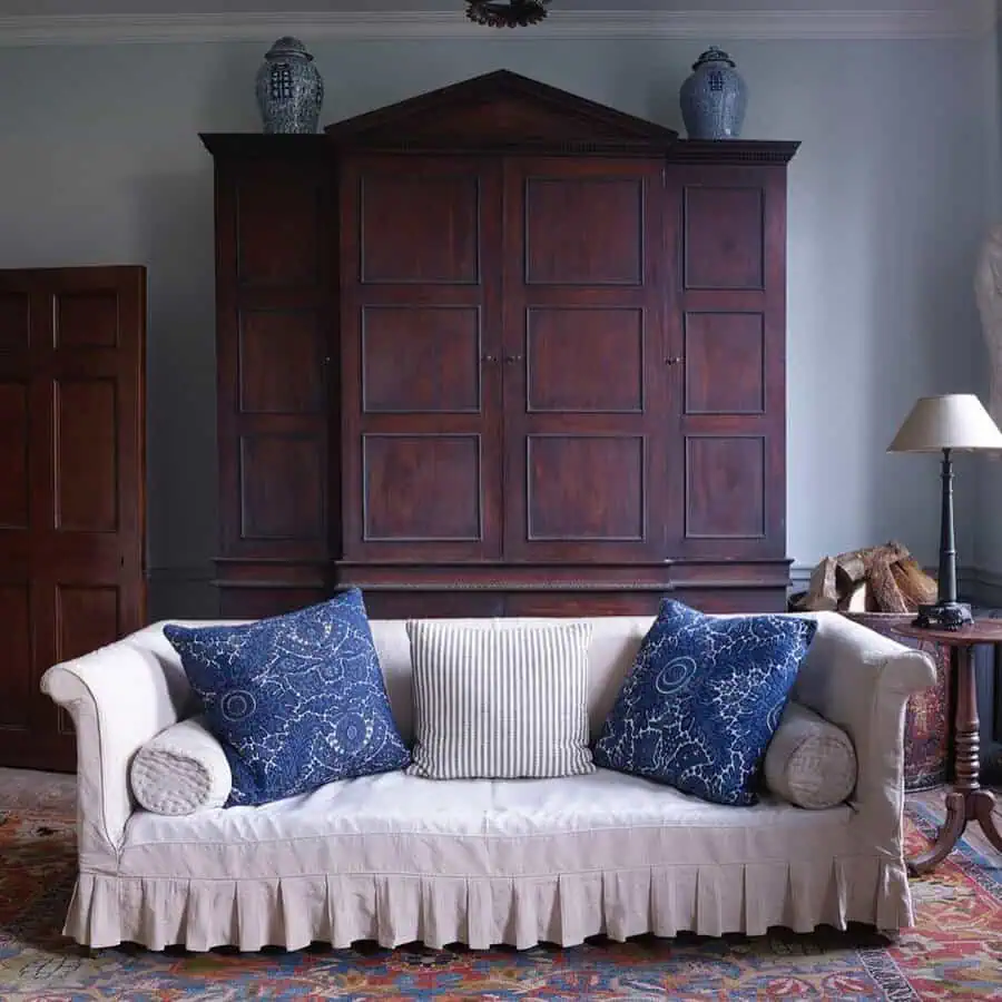 Serene sitting room with a tall mahogany armoire, pale blue walls, a slipcovered sofa, and indigo pillows for a refined Georgian touch.