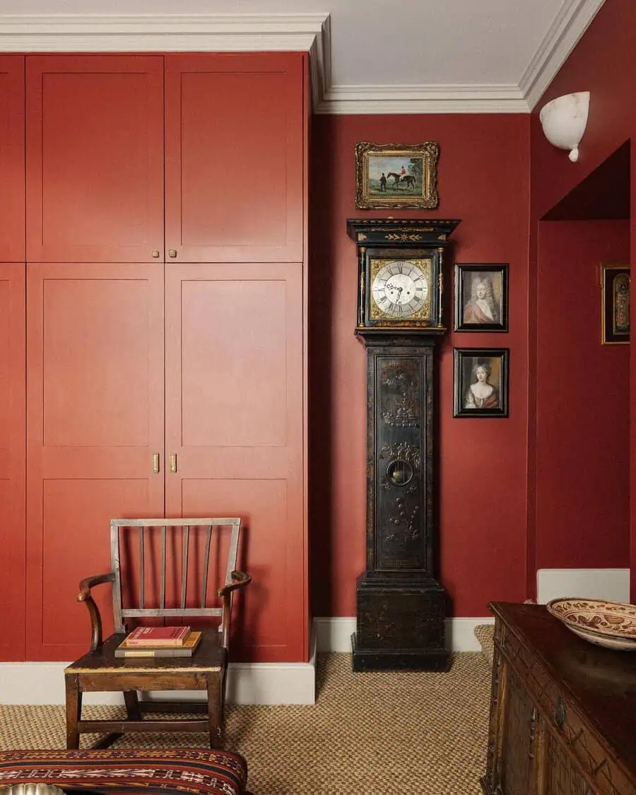 Painted grandfather clock displayed against red walls, framed by vintage portraits and a modest wooden chair for a classic look.