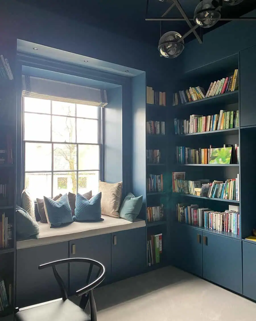 Cozy reading nook with built-in shelves, blue walls, a comfy window seat, and soft lighting for a perfect reading space.