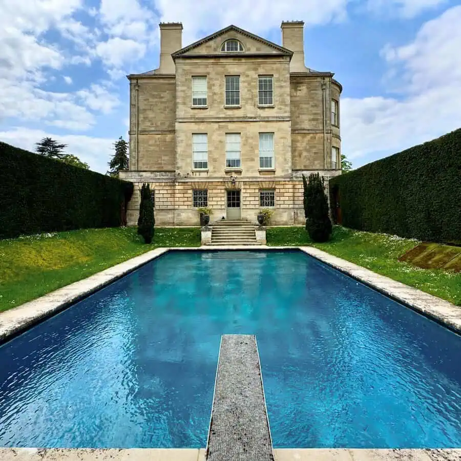 Large rectangular pool leading to a grand Georgian house, flanked by tall hedges and stone steps in a formal garden setting.