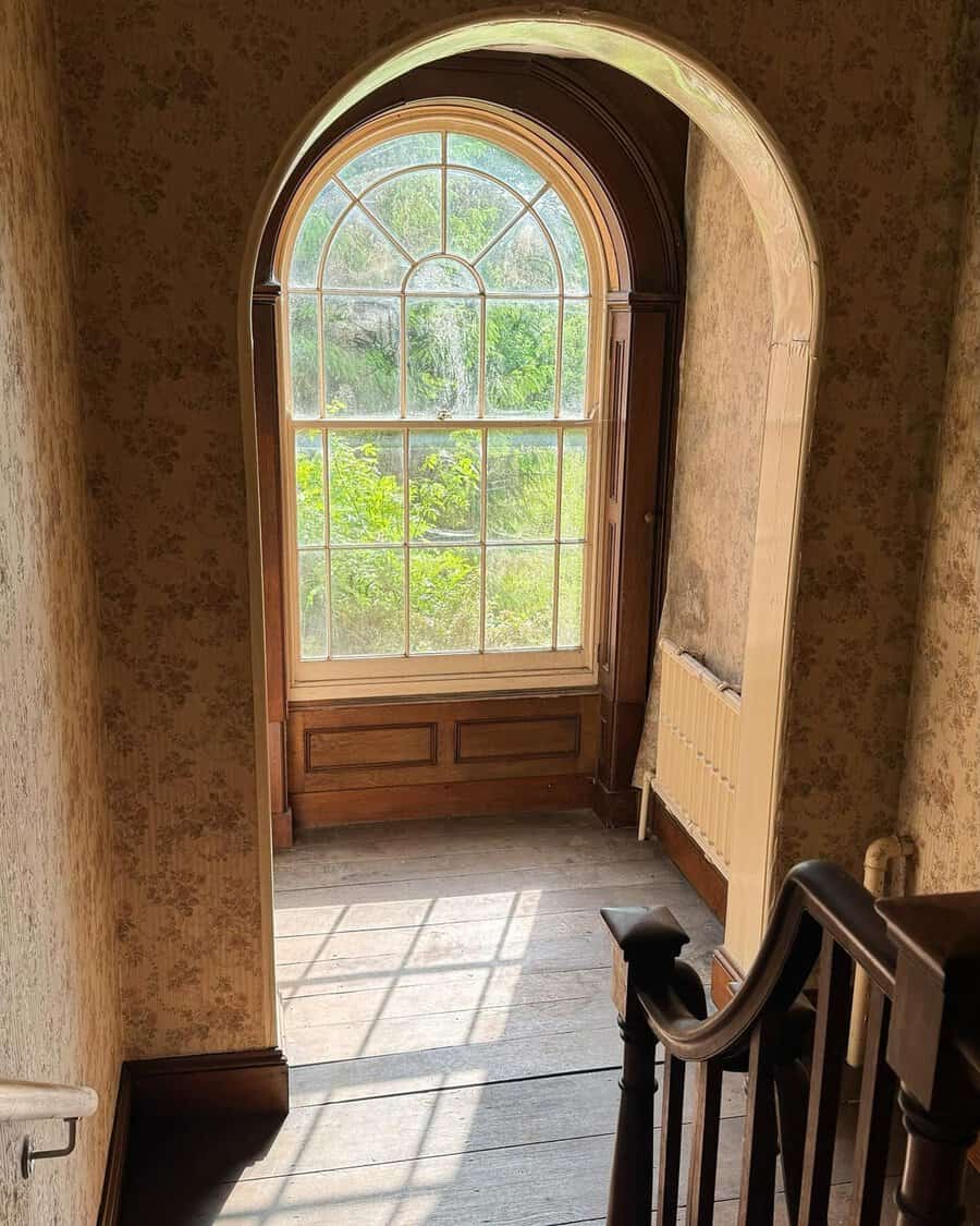 Georgian stairwell with arched window, wood flooring, and floral wallpaper bathed in gentle sunlight.