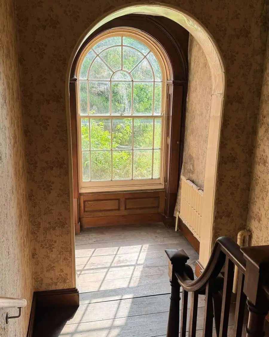 Georgian stairwell with arched window, wood flooring, and floral wallpaper bathed in gentle sunlight.